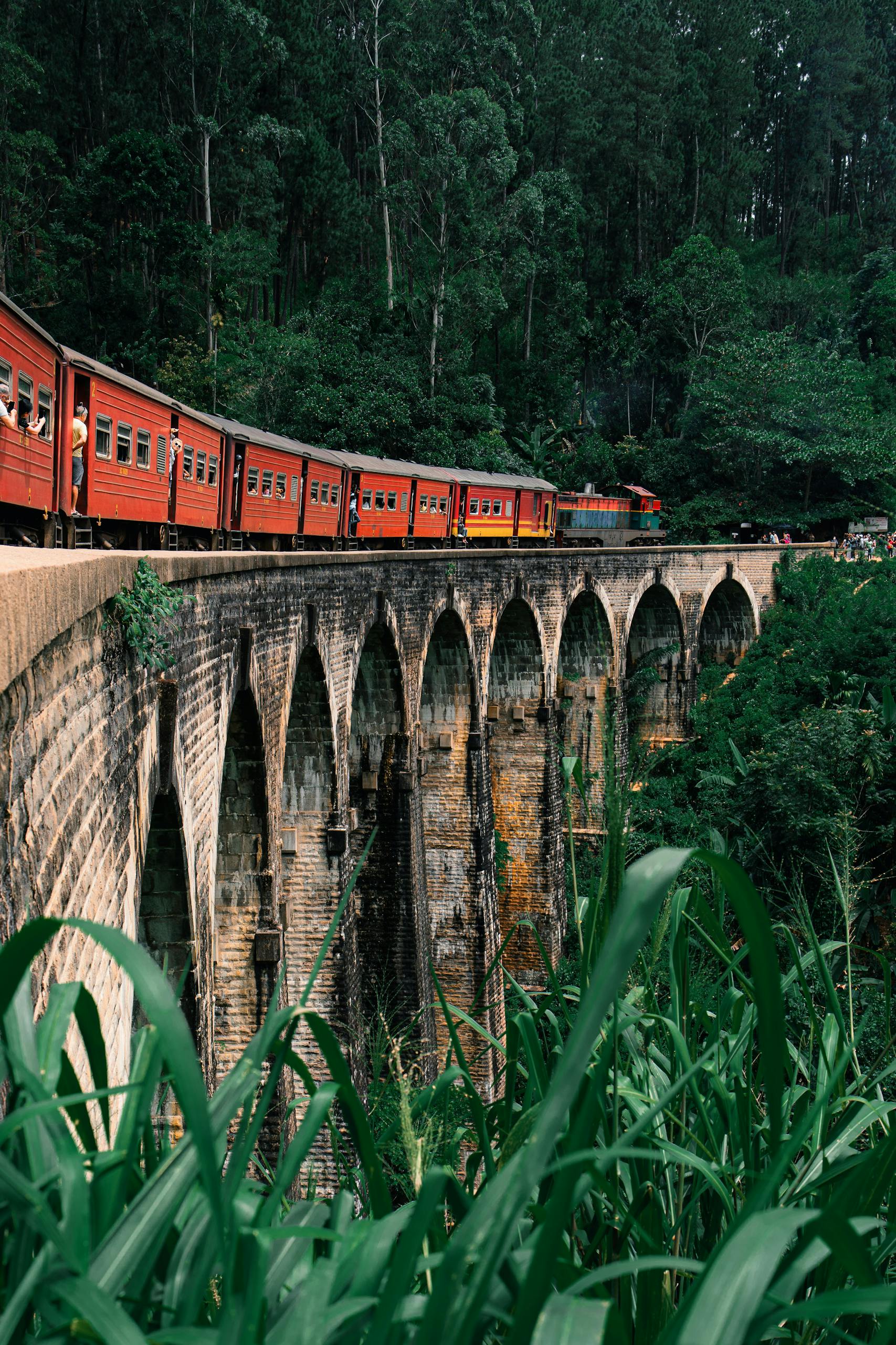 A vibrant red train travels across the historic Nine Arches Bridge in Ella, Sri Lanka, surrounded by lush greenery.
